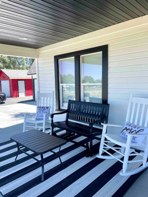 A porch with two rocking chairs, a black bench, and a coffee table on a striped rug, with a red shed in the background.